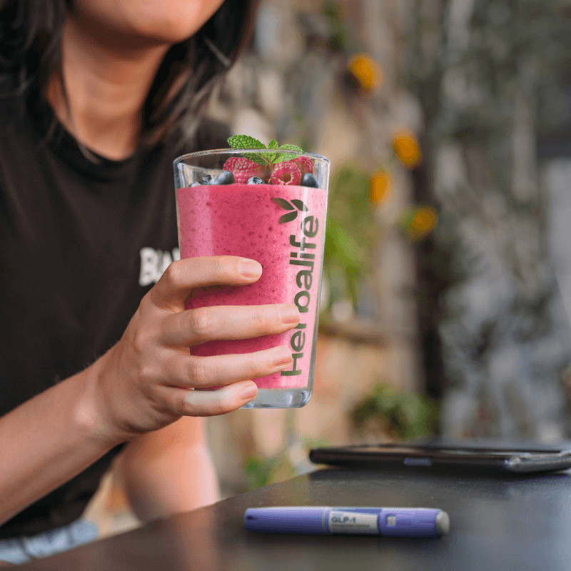 A person holding a Herbalife protein shake glass filled with berry smoothie, showcasing the NaturalVive health drink.